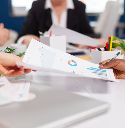 Confident company manager giving working tasks to diverse teamworkers analysing paperwork with graphs sitting in start up office. Colleagues working in office at conference table in startup management company.